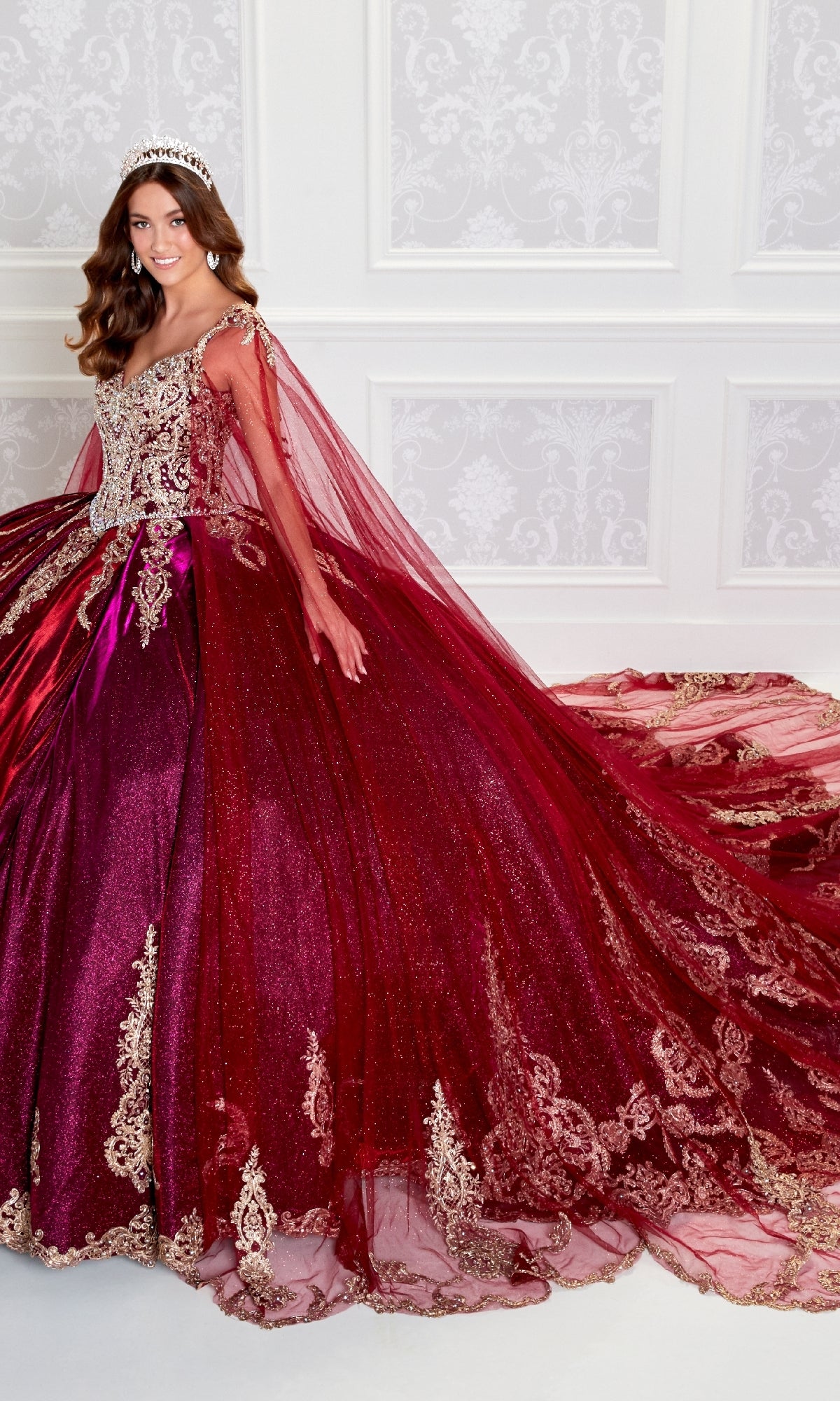 A woman stands against an ornate white wall wearing the Quinceanera Dress PR22141 By Princesa—a sparkling burgundy ball gown with gold embroidery, corset back, long train, and a tiara.